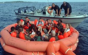 Auf dem Bild sieht man ein oranges Rettungsboot auf dem Meer, welches 9 Menschen beinhaltet. Dahinter ist ein weißes Motorboot mit zwei Männern, die zu den anderen herunterschauen.